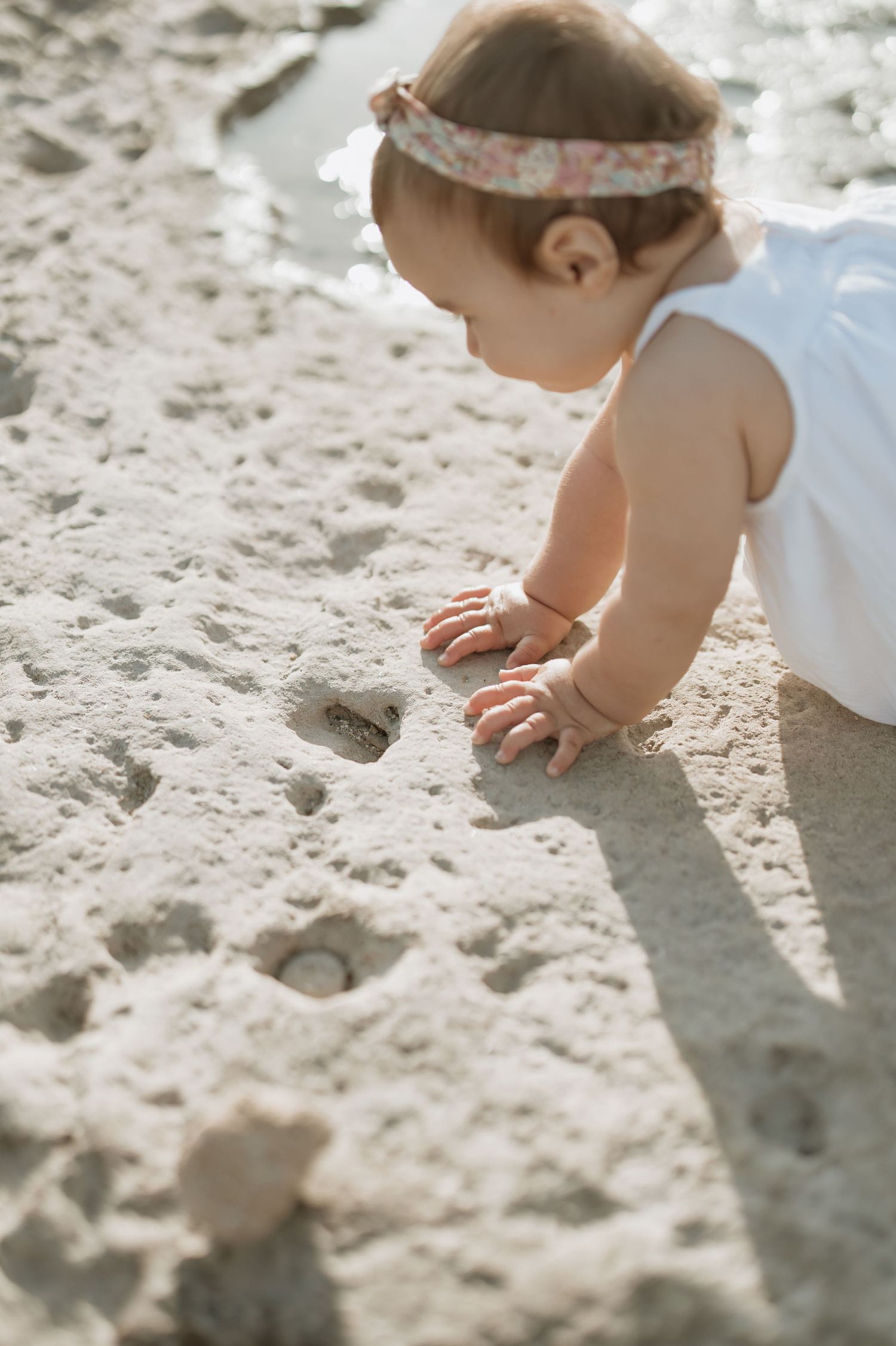 A baby in white dress explores the sandy beach surface.