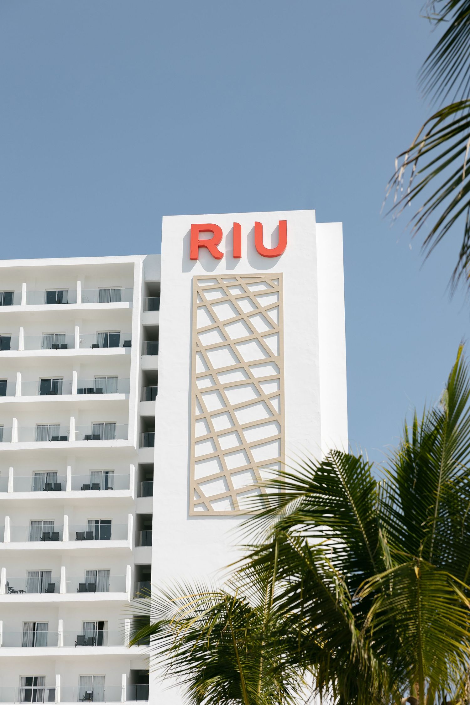 White RIU hotel building with geometric design and palm trees in foreground.