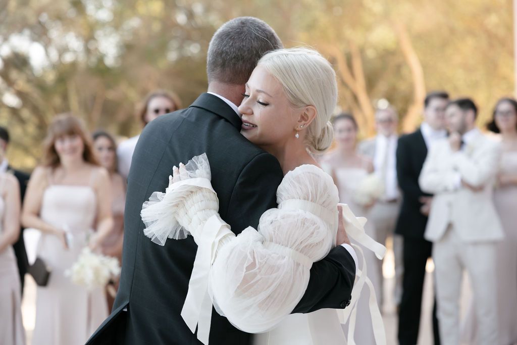 A tender embrace during a wedding dance with guests watching in soft daylight.