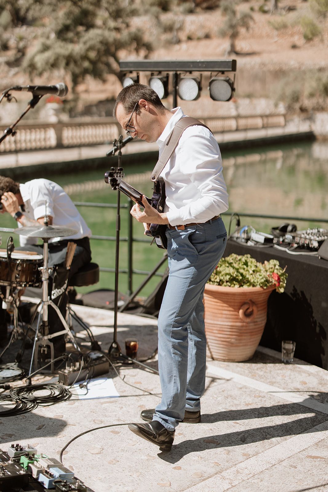 Band member plays guitar while performing at an outdoor venue with stage lighting and terrace setting.