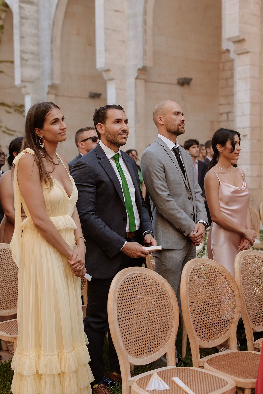 Wedding guests in formal attire stand beside elegant cane-back chairs during an outdoor ceremony.