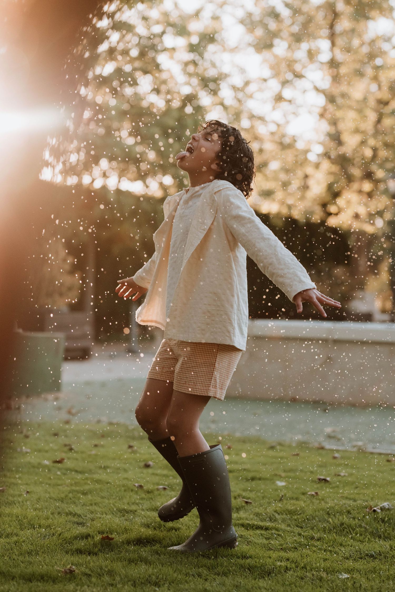 Silhouetted figure dances joyfully in raindrops at golden hour wearing a white sweater and rain boots on grassy lawn.