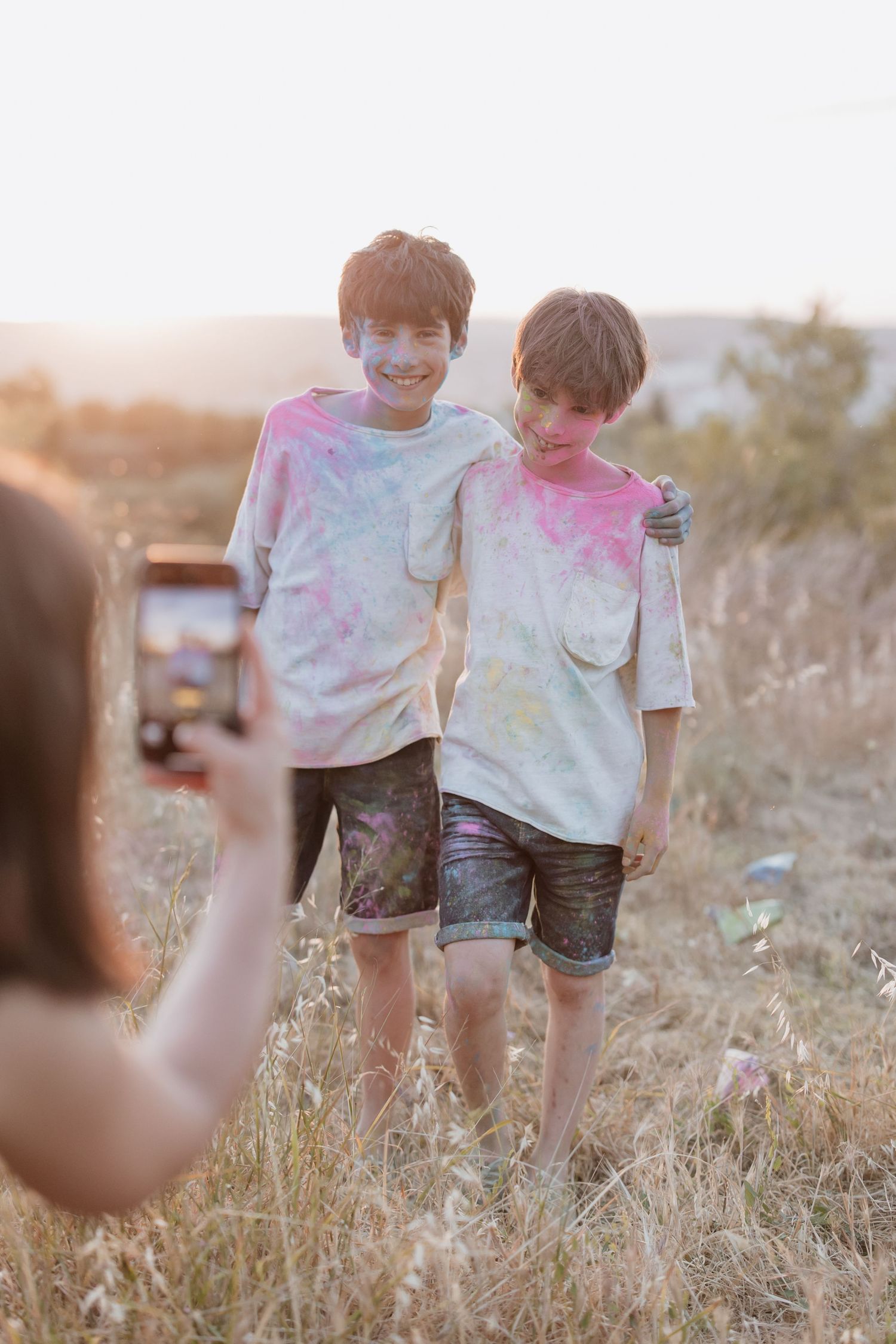 Two children wearing colorful tie dye shirts pose together in a field at sunset while someone takes their photo.