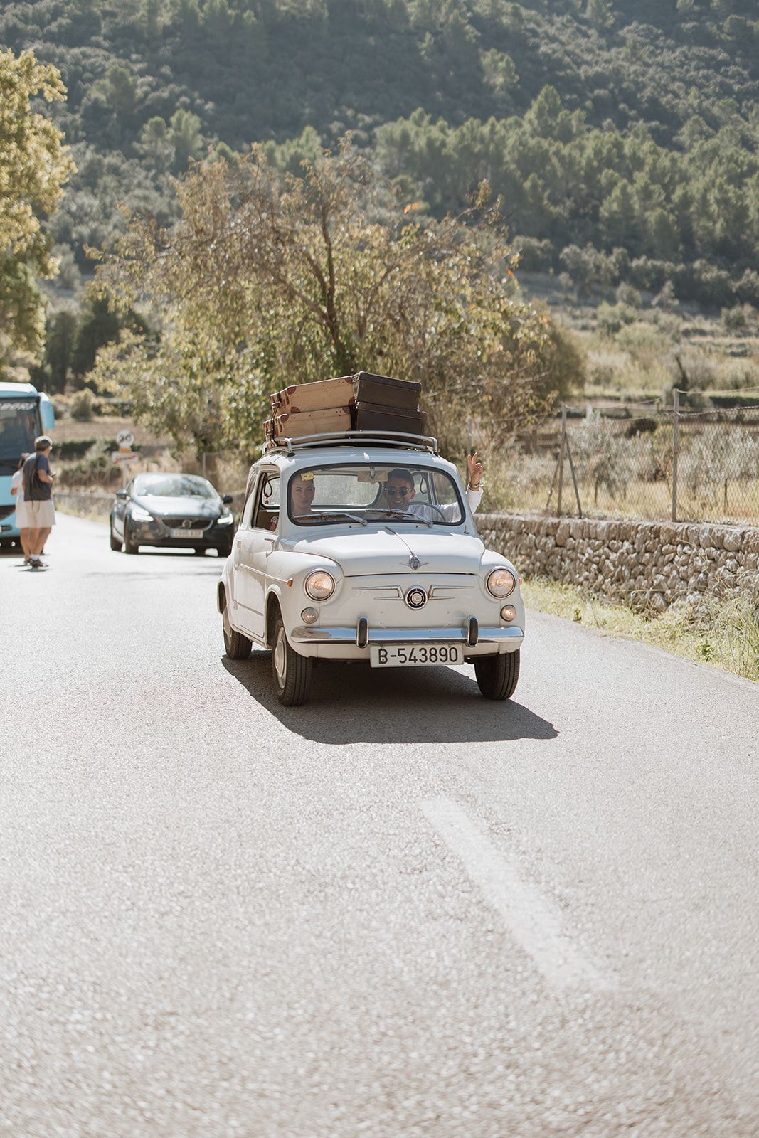 Classic Fiat 500 car driving on rural road with luggage on roof.