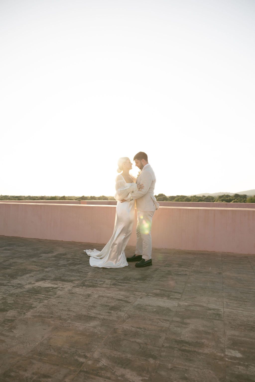 Newlyweds embrace during golden hour on rooftop with lens flare.