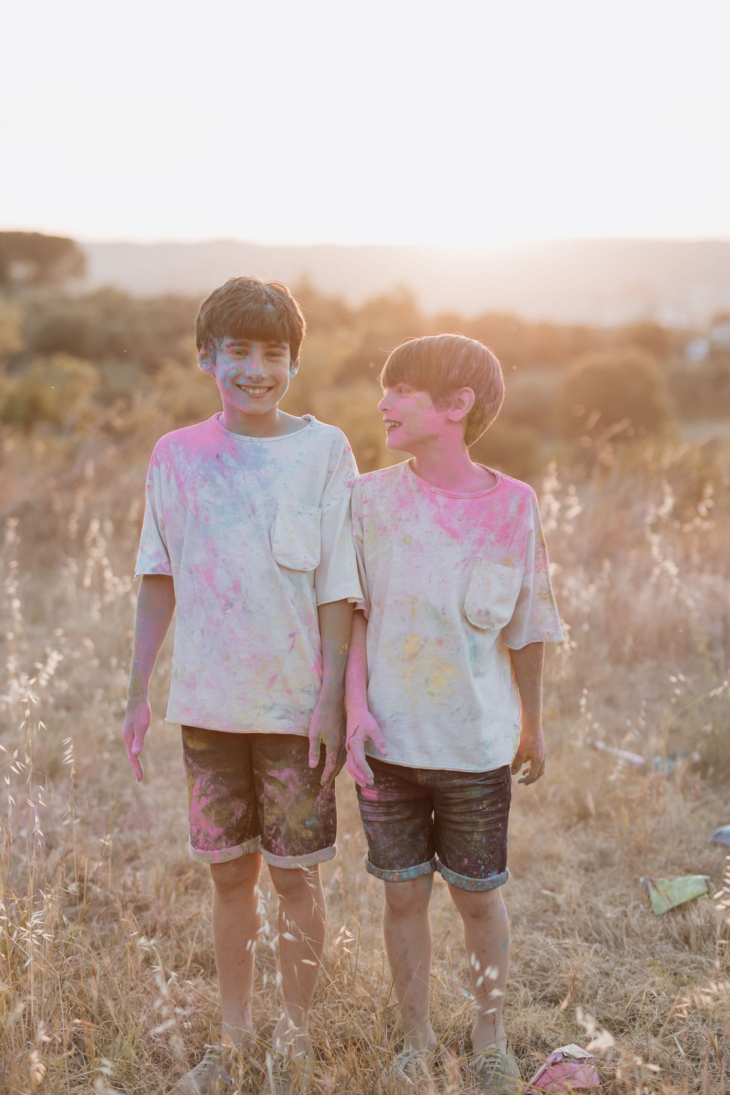 Two children wearing colorful tie-dyed t-shirts stand together in a sunlit field of dry grass at golden hour.