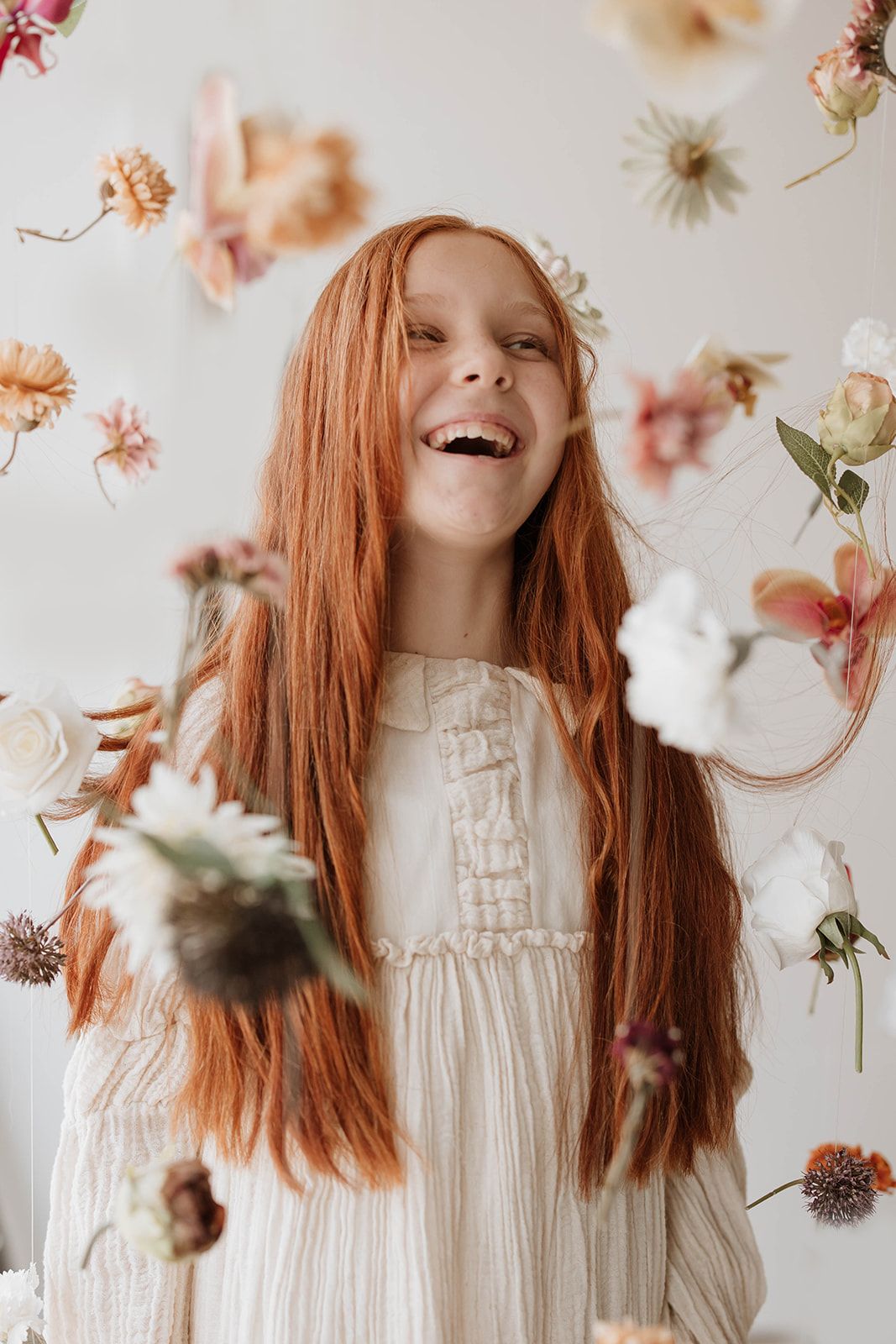 Natural light portrait with flowing red hair and white dress among floating dried flowers.