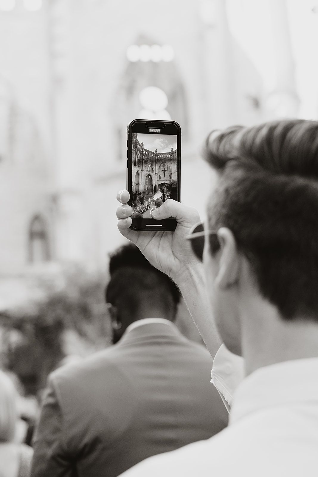 A guest captures the wedding ceremony on their phone in black and white.