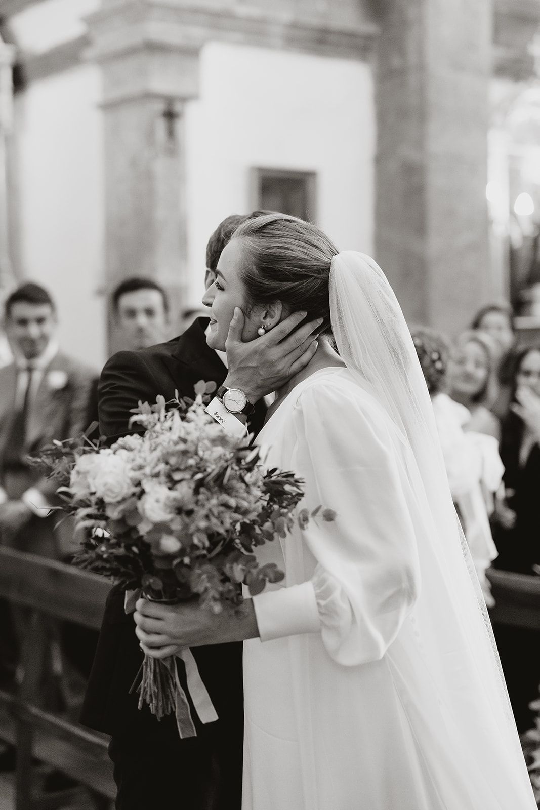 Intimate black and white wedding moment captured in a church with the bride holding an elegant floral bouquet.