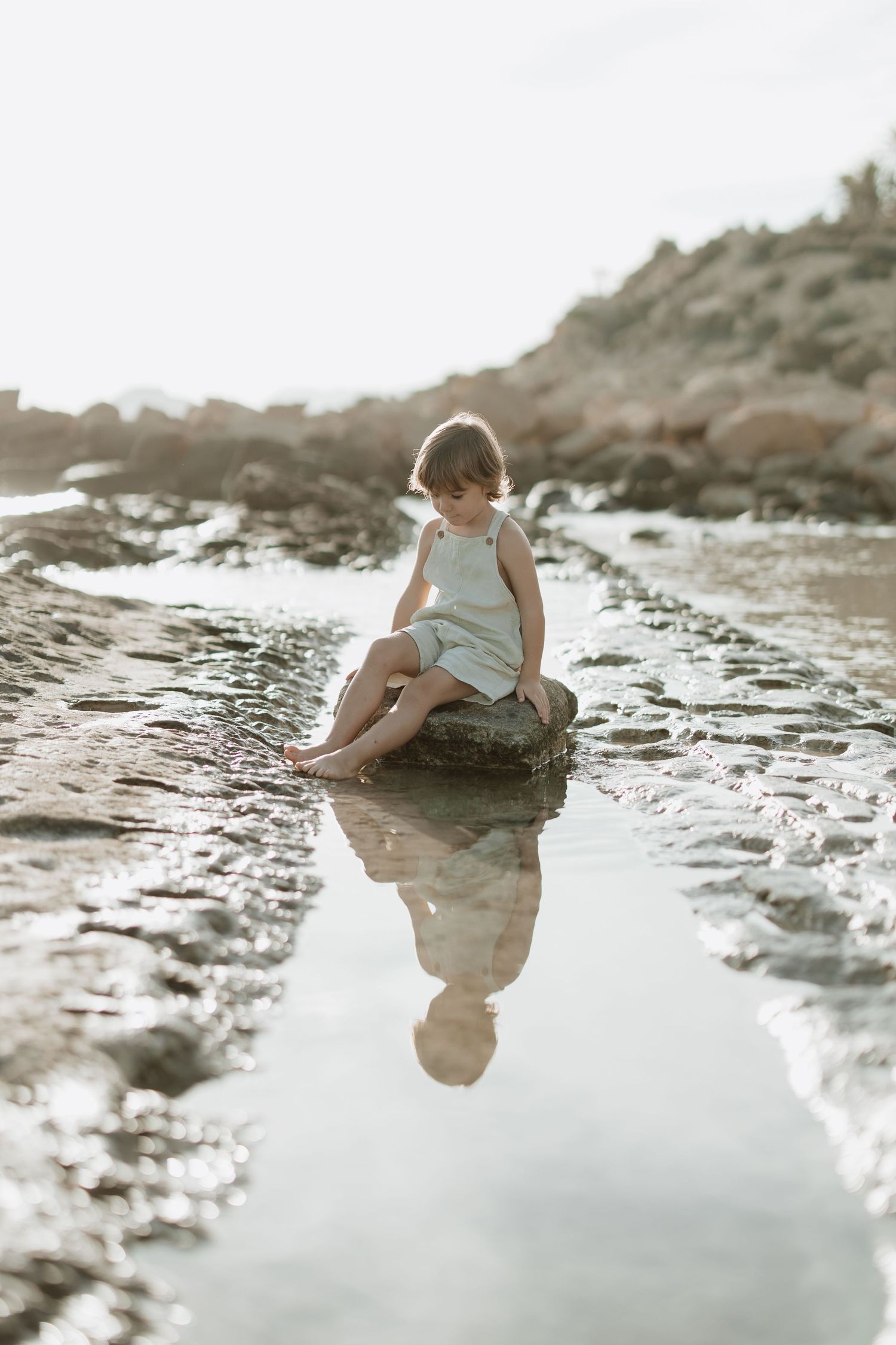 Child sits on a rock at the beach with reflection in shallow water at sunset.