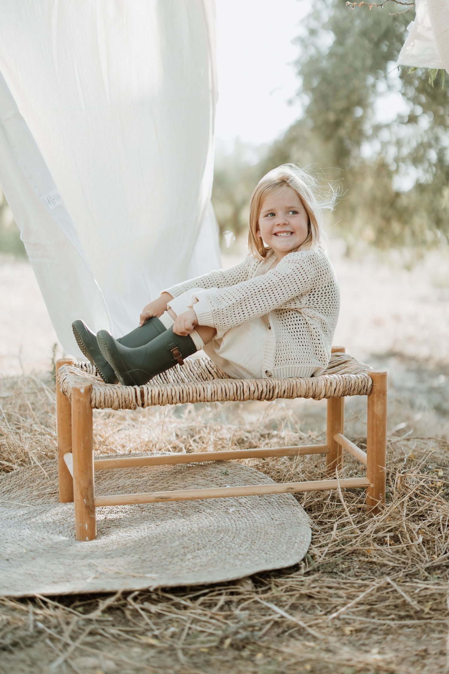 Child in cream knit sweater and green boots sitting on rustic woven bench outdoors with white fabric backdrop.