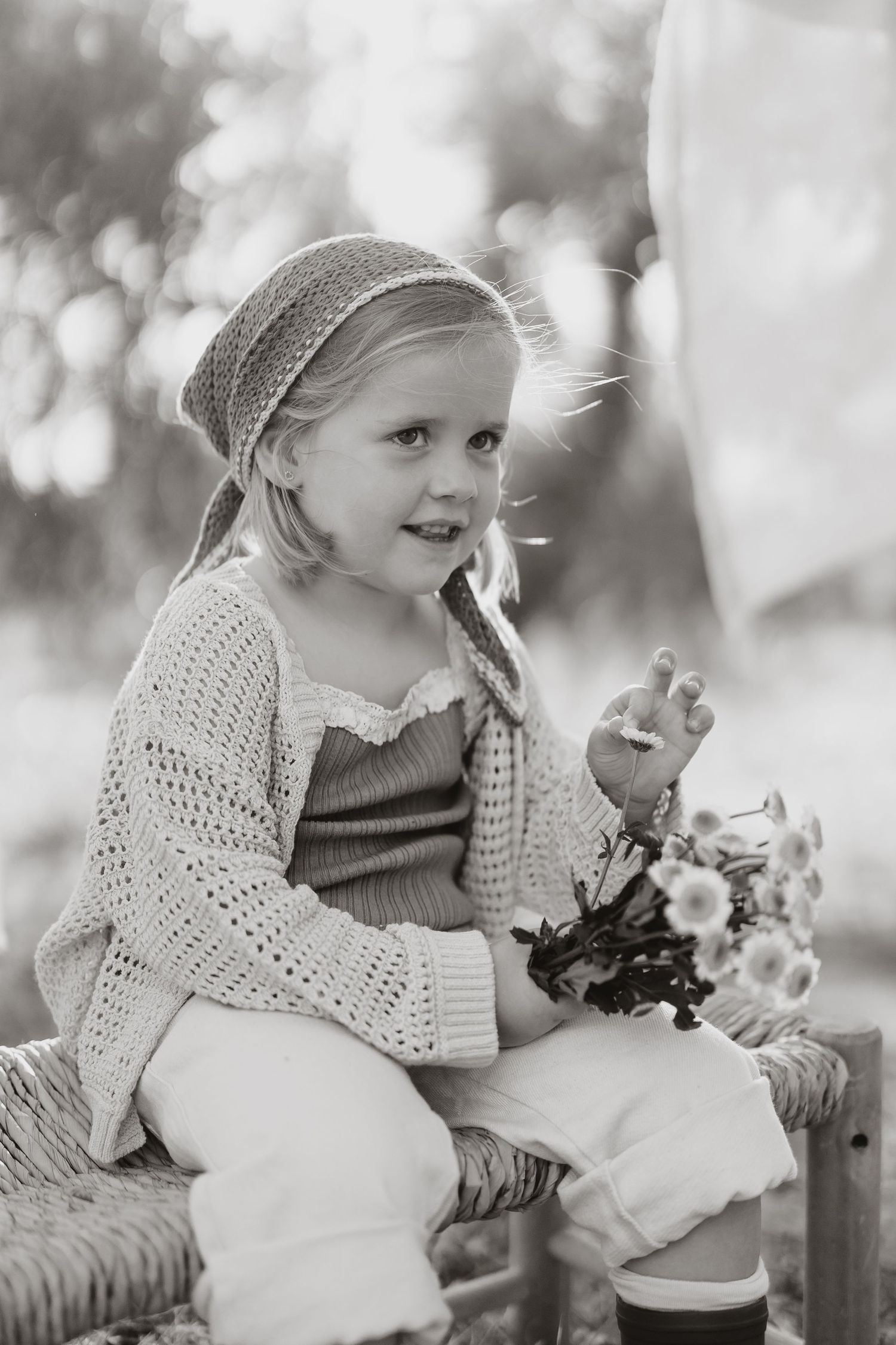 Black and white portrait of child in headscarf and sweater holding flowers on rustic bench.