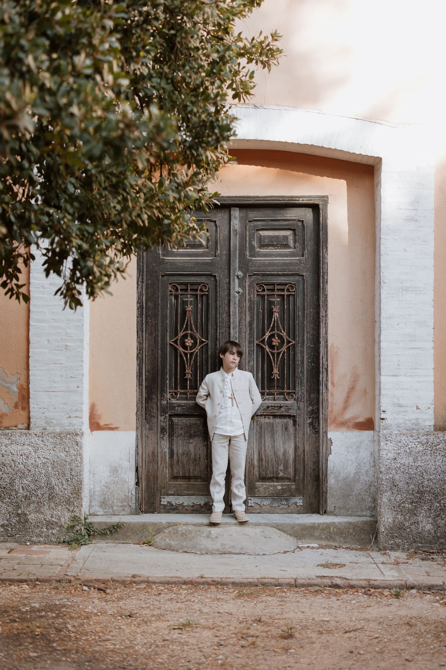 A person in light colored clothing stands in front of an ornate vintage double door entrance with tree branches overhead.