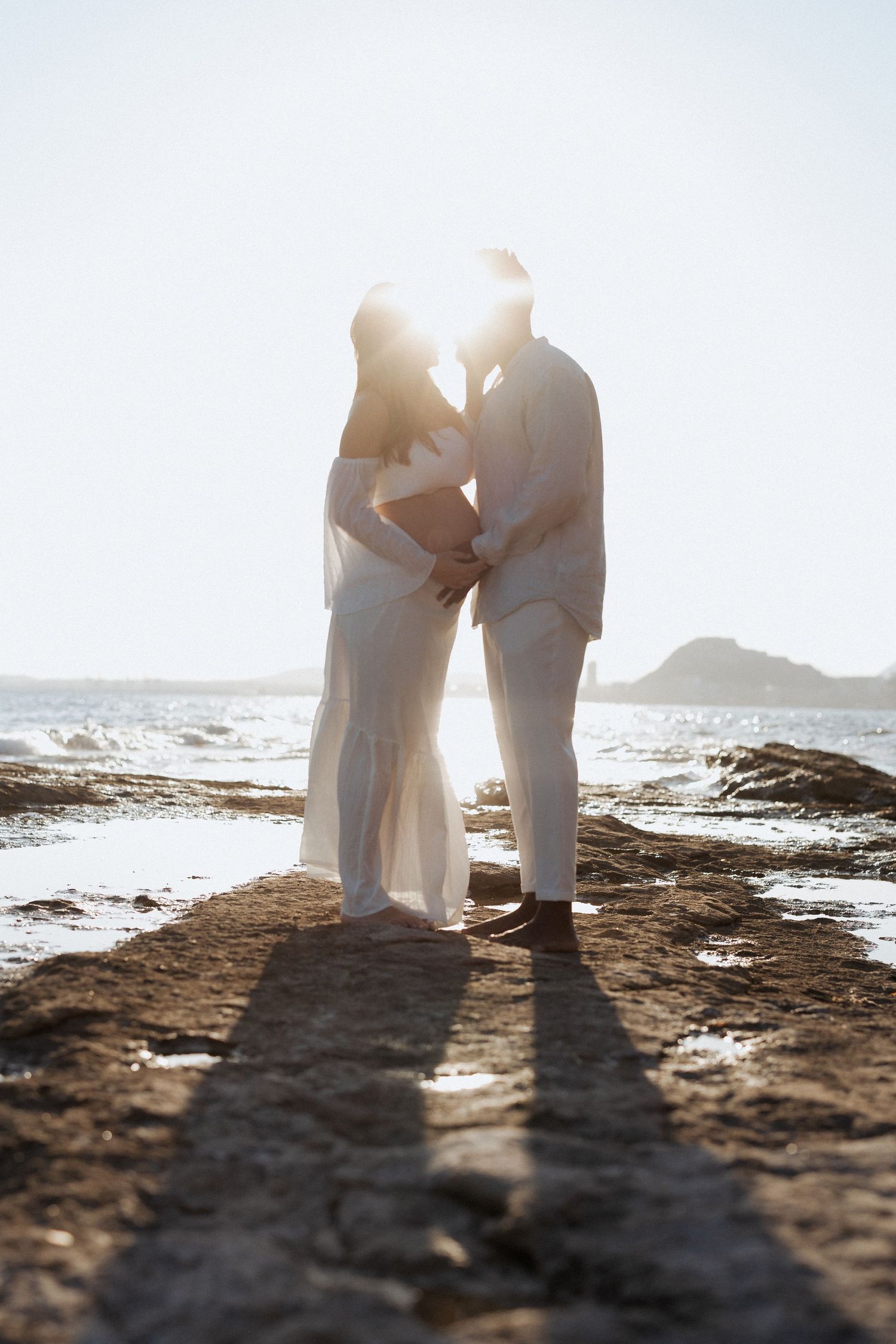 Silhouetted couple in white clothing stands together on rocky beach at sunset casting long shadows on sand.