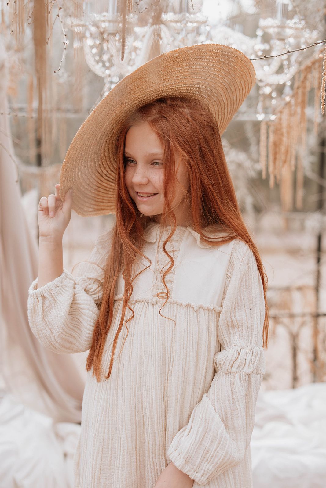 Soft portrait with straw sunhat and white dress against crystal and fabric decorations.