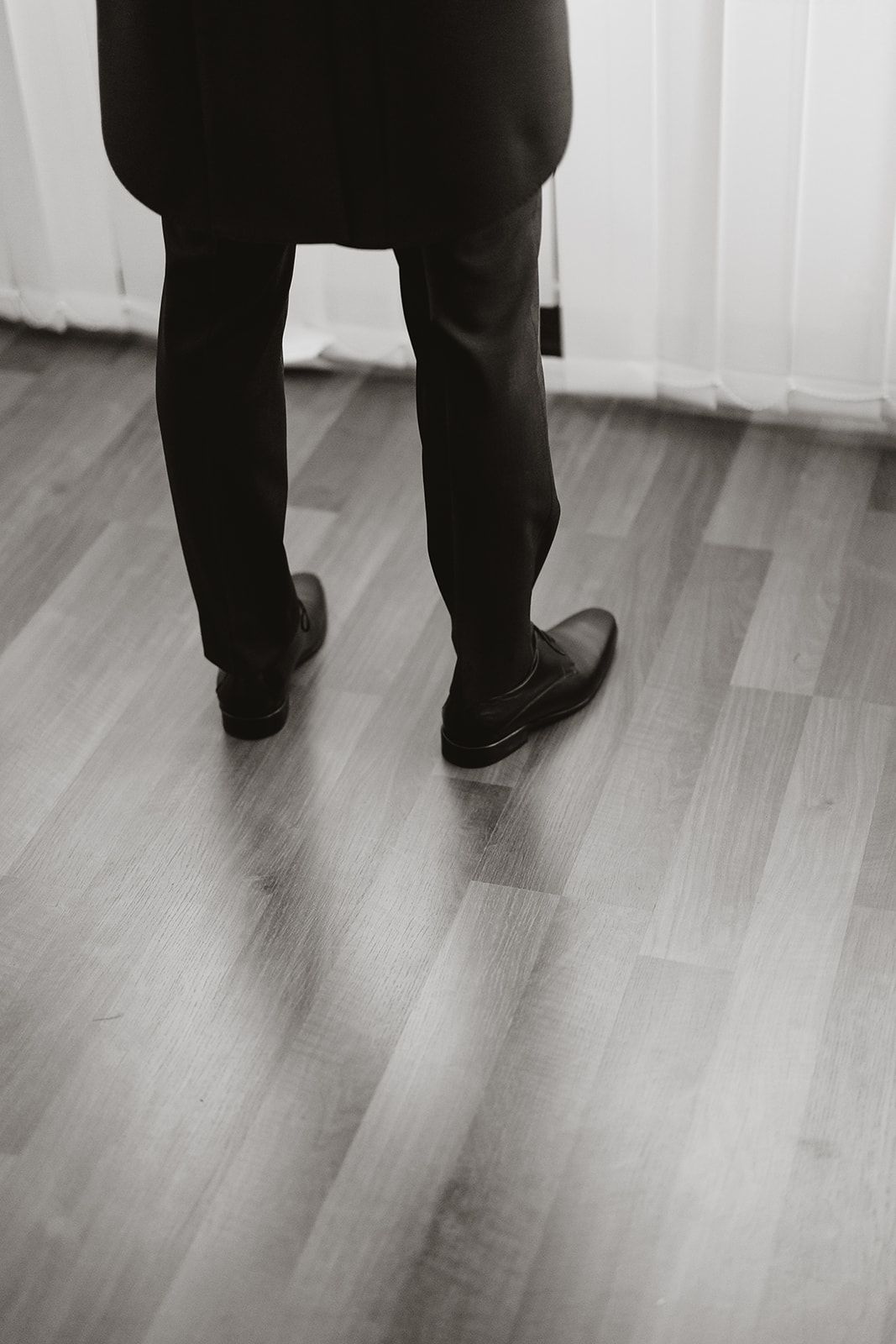 Black and white photo of dress shoes and suit pants on hardwood floor.