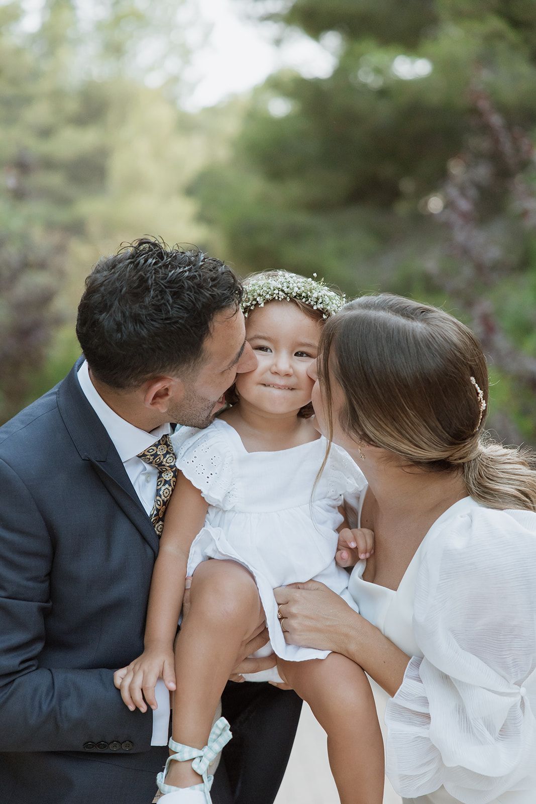 Sweet family moment shared between parents and young flower girl at outdoor wedding.