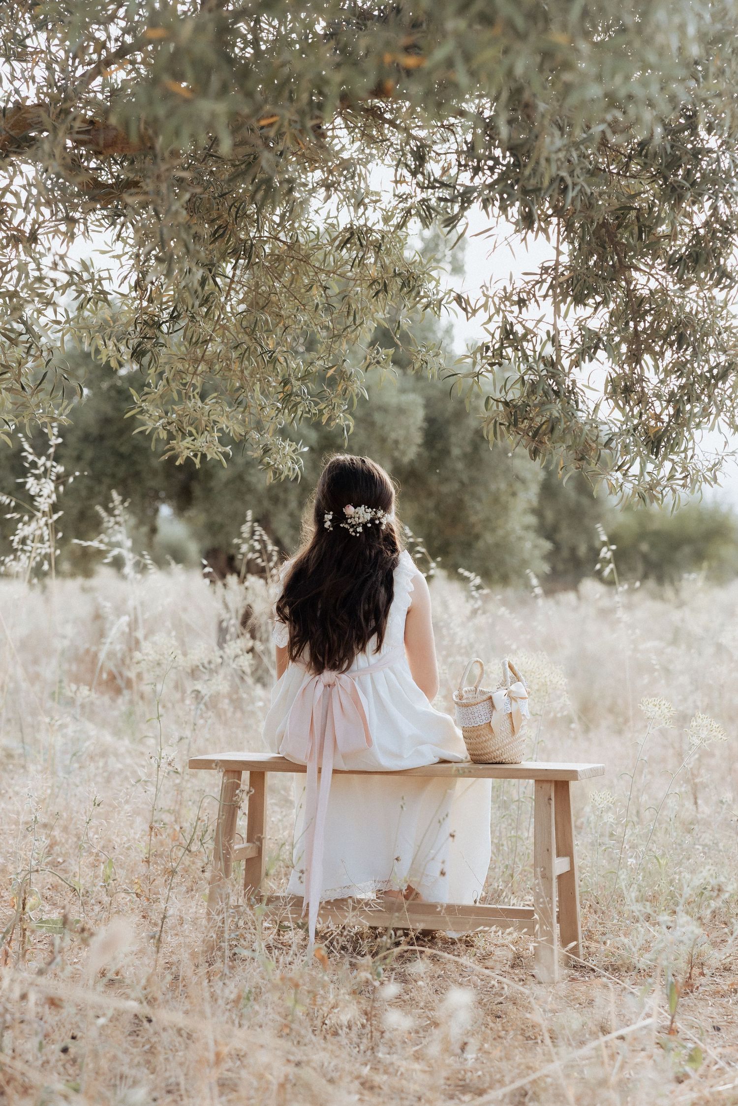 Young woman in white dress with floral hair accessory sitting on wooden bench in a serene meadow under olive tree.