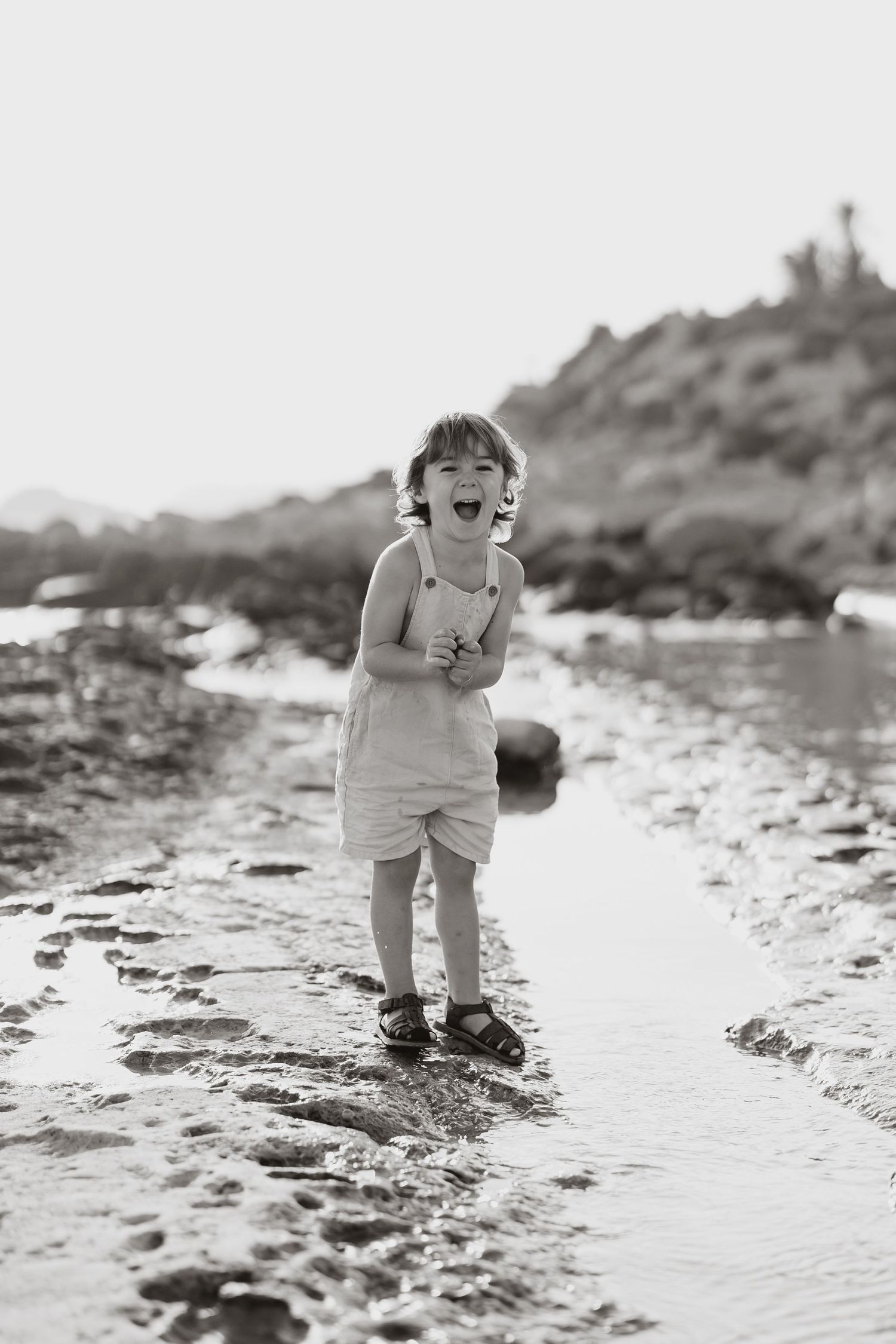 A small child plays joyfully in shallow water along a rocky shoreline.