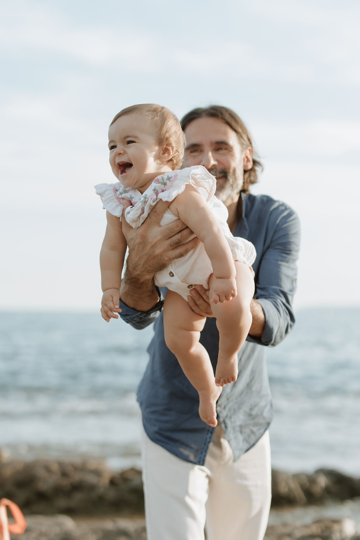 A happy baby in a white outfit being held up against an ocean backdrop.