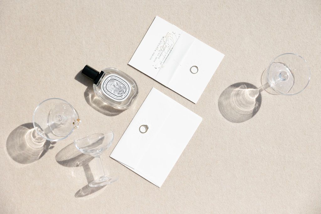 Minimalist flatlay with perfume bottle, white cards and champagne glasses on beige surface.