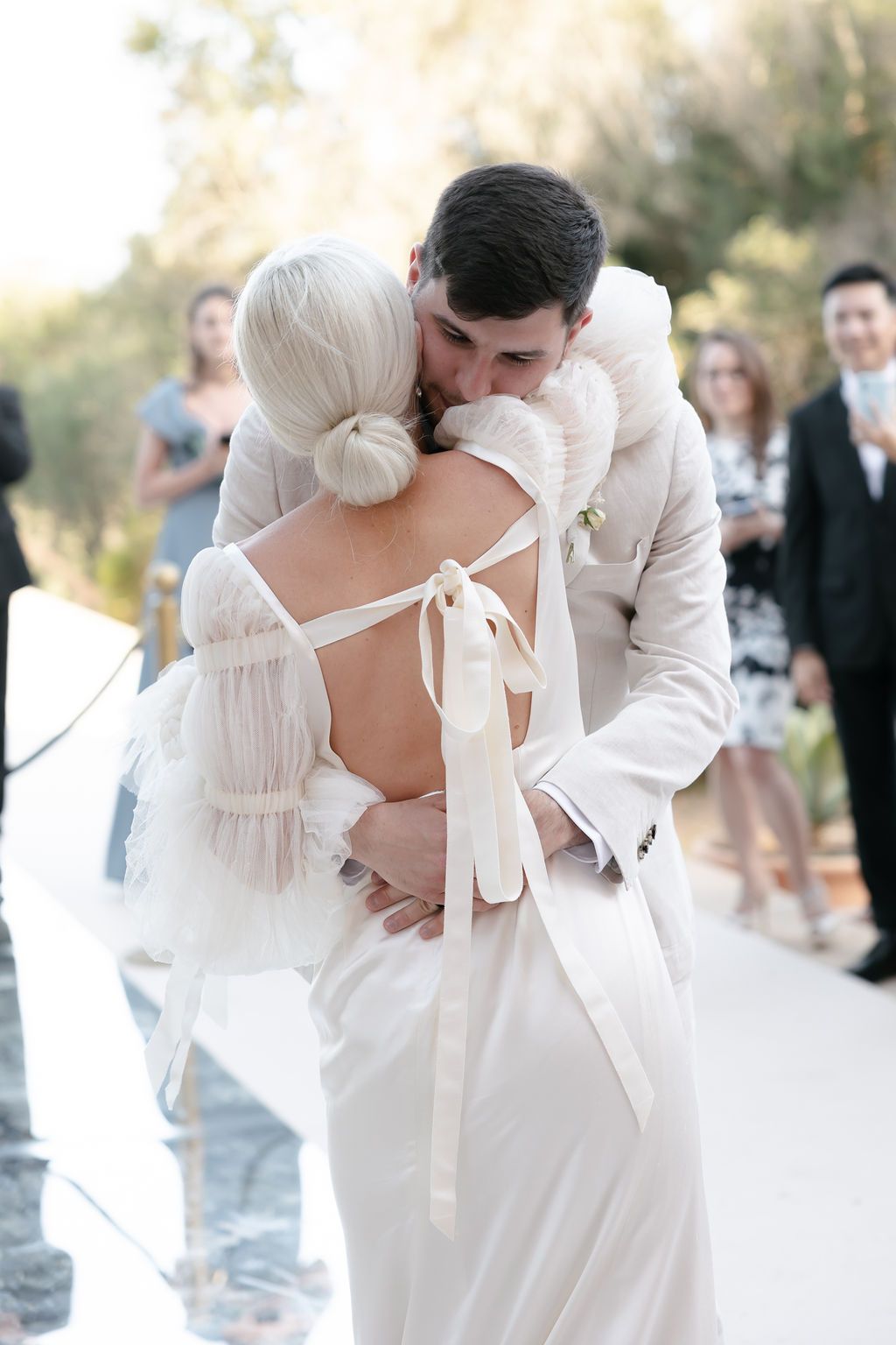 A romantic wedding dance embrace shows the couple in white with guests looking on in the background.