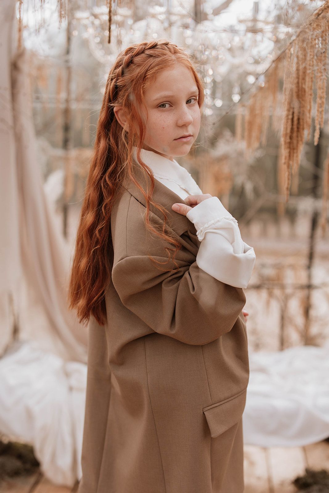 Dreamy portrait in brown coat with intricately braided hair in rustic greenhouse setting.
