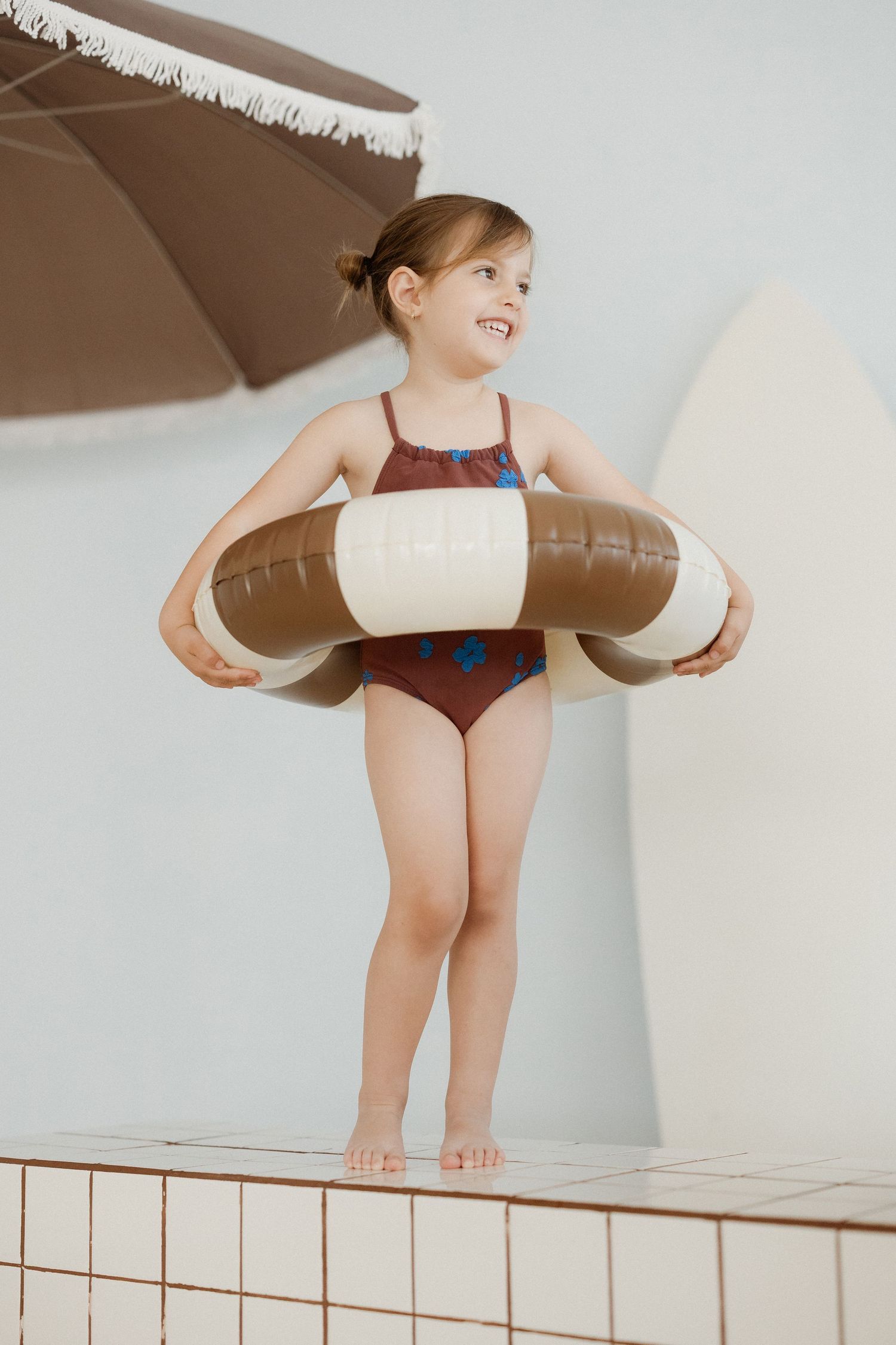 Young girl in floral swimsuit holding brown inflatable pool ring, standing poolside near beach umbrella.