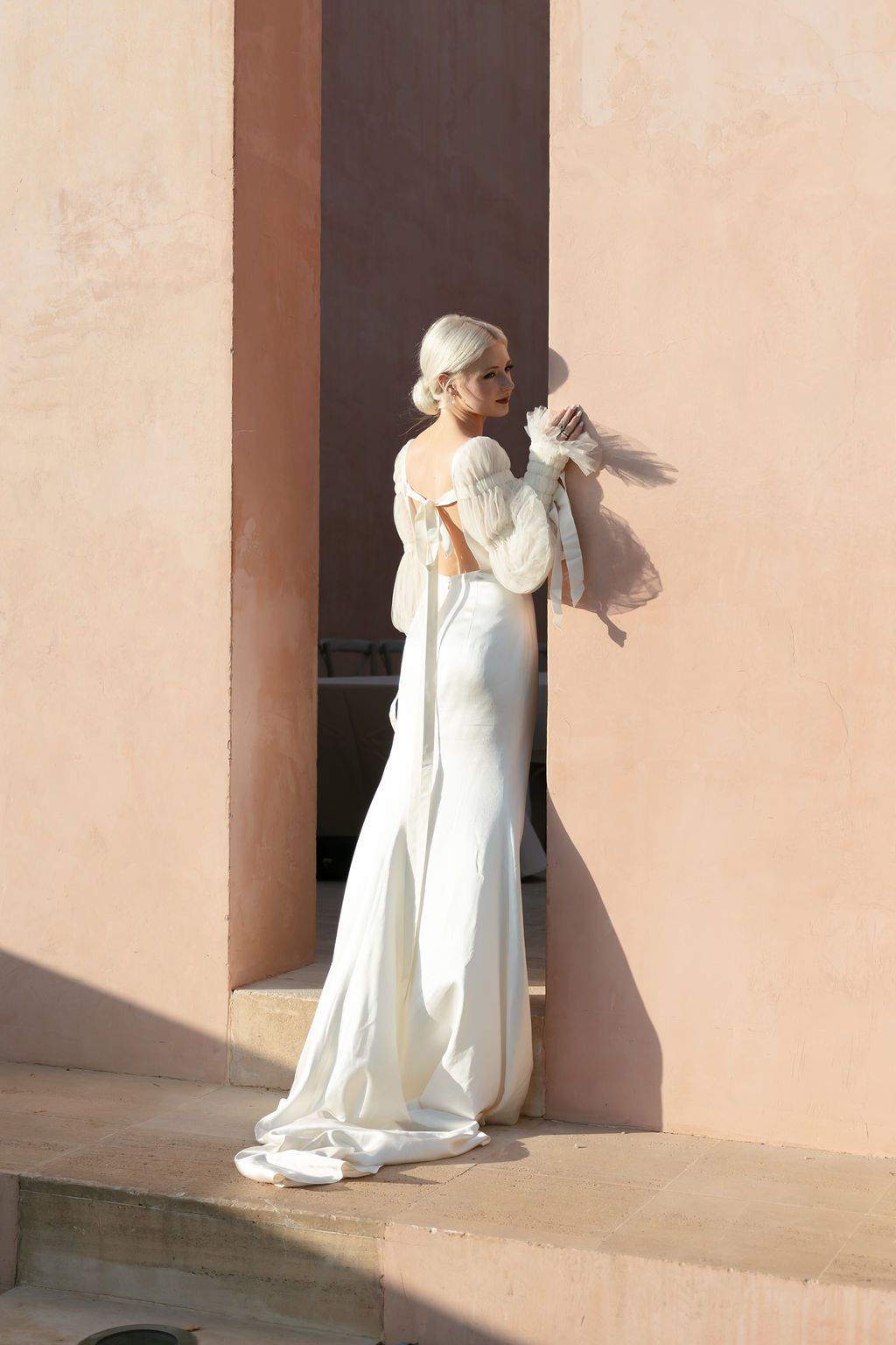 Elegant model in flowing white gown poses gracefully against peachy wall in natural lighting.