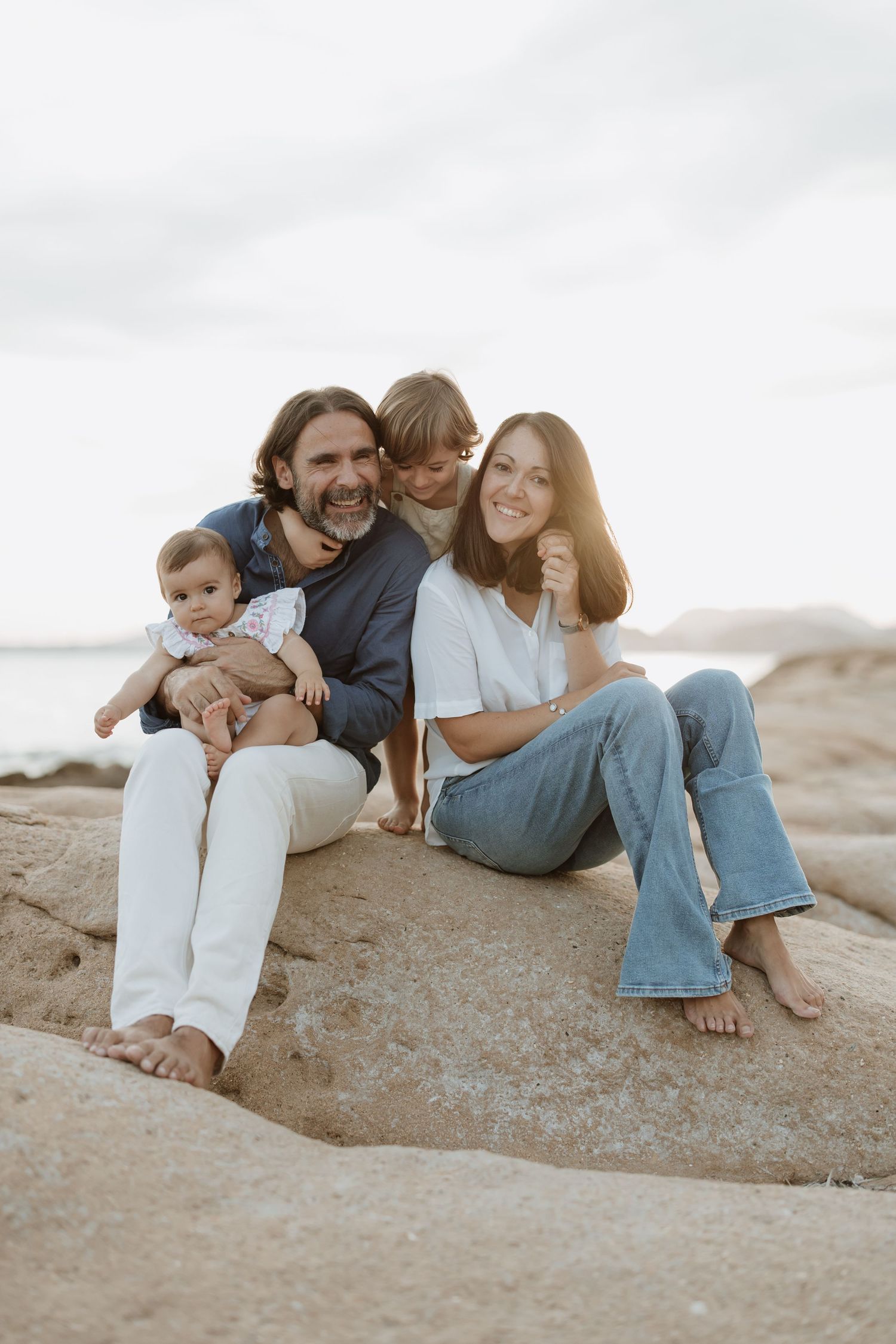 Family relaxes together on coastal rocks during sunset photoshoot at beach.