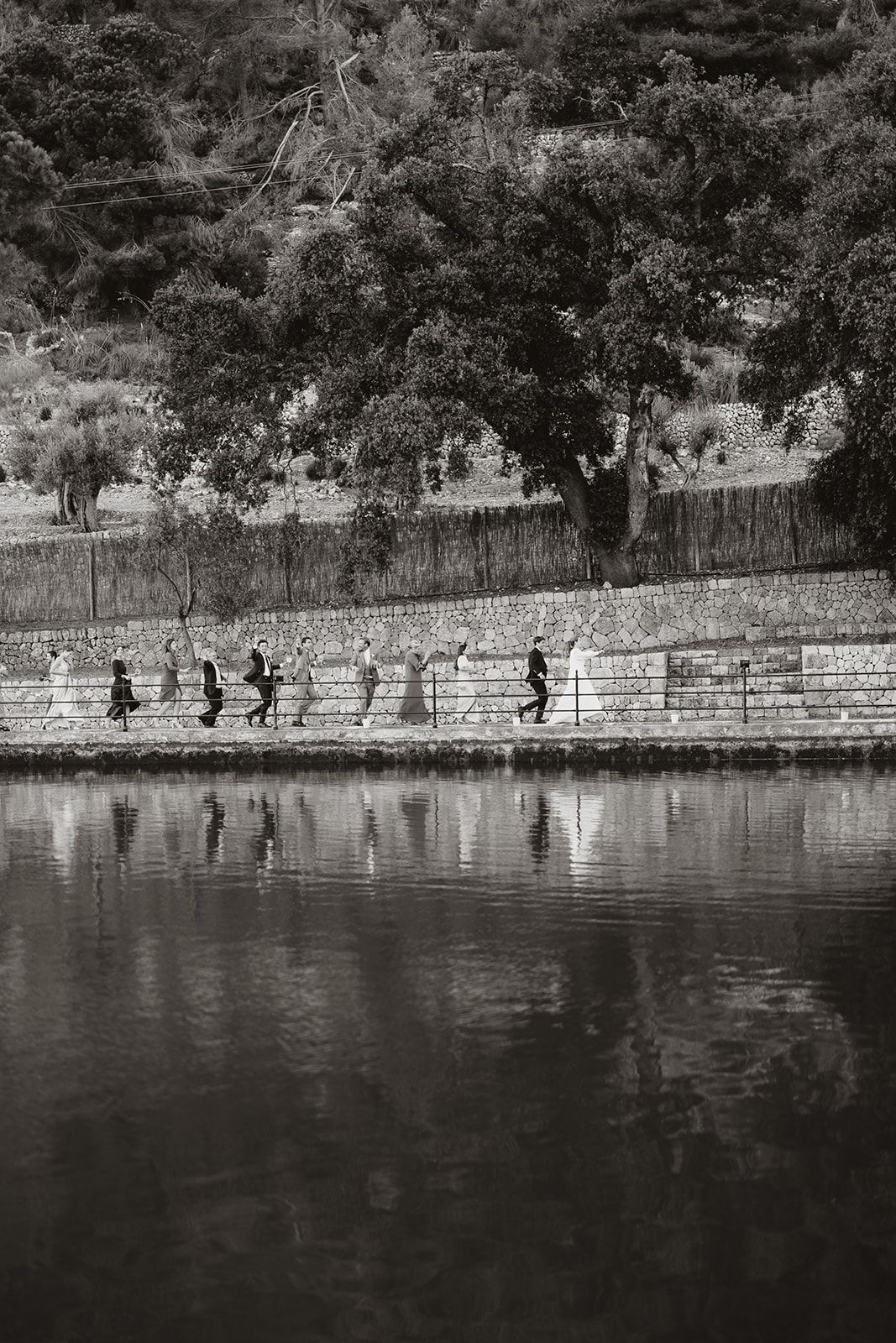 Black and white image of silhouettes walking along a waterfront wall with reflections in the water.