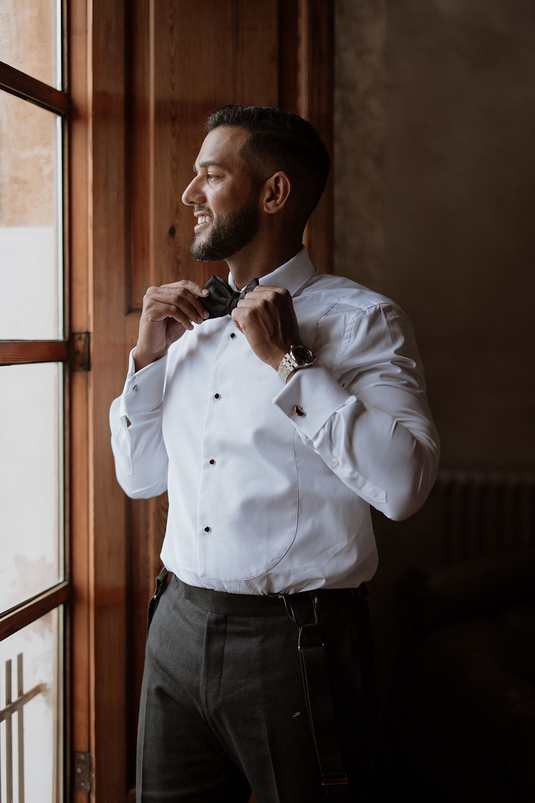 A person in formal attire adjusts their bow tie while standing by a window.
