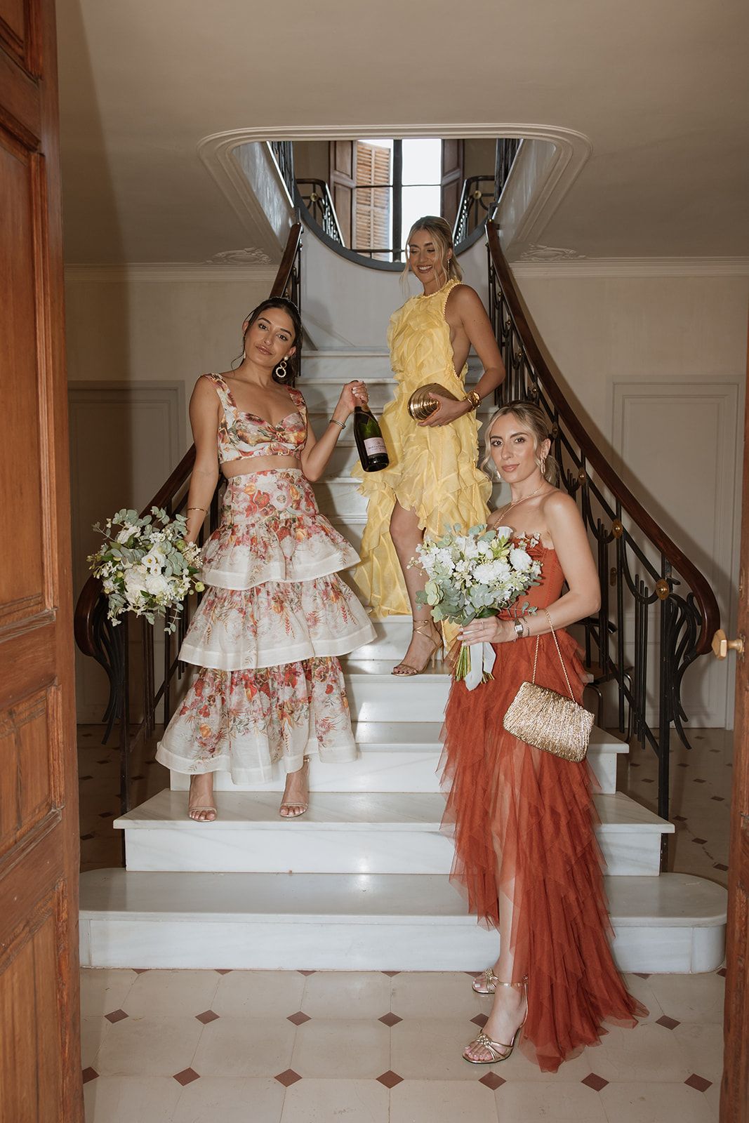 Three women in elegant dresses pose with champagne and bouquets on an elegant white staircase for a formal event.