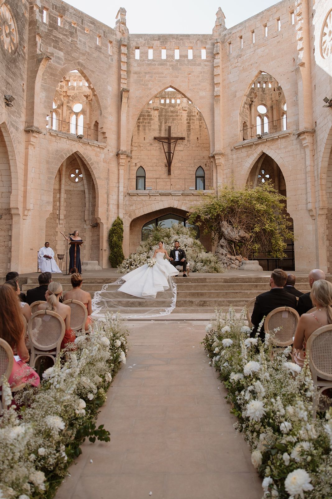 An outdoor wedding ceremony features white floral arrangements and cane chairs against gothic architecture.