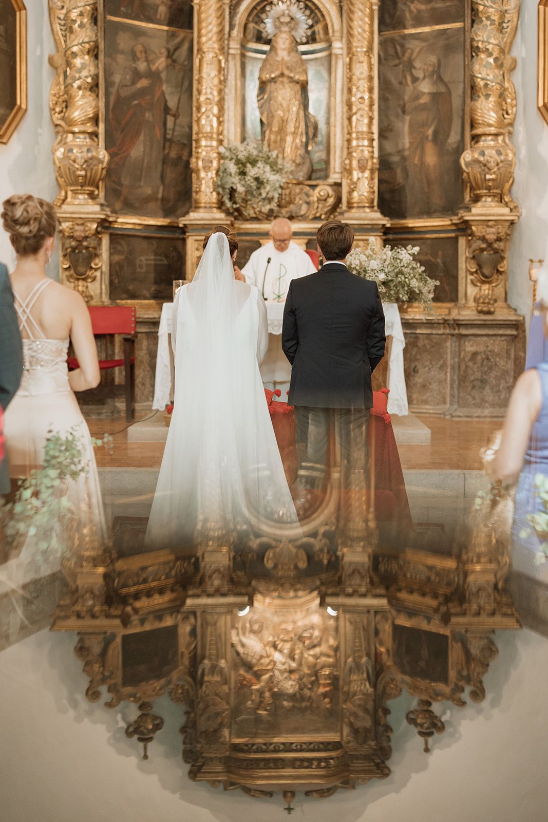 Wedding couple stands before an ornate golden altar with religious artwork and decorative floral arrangements.