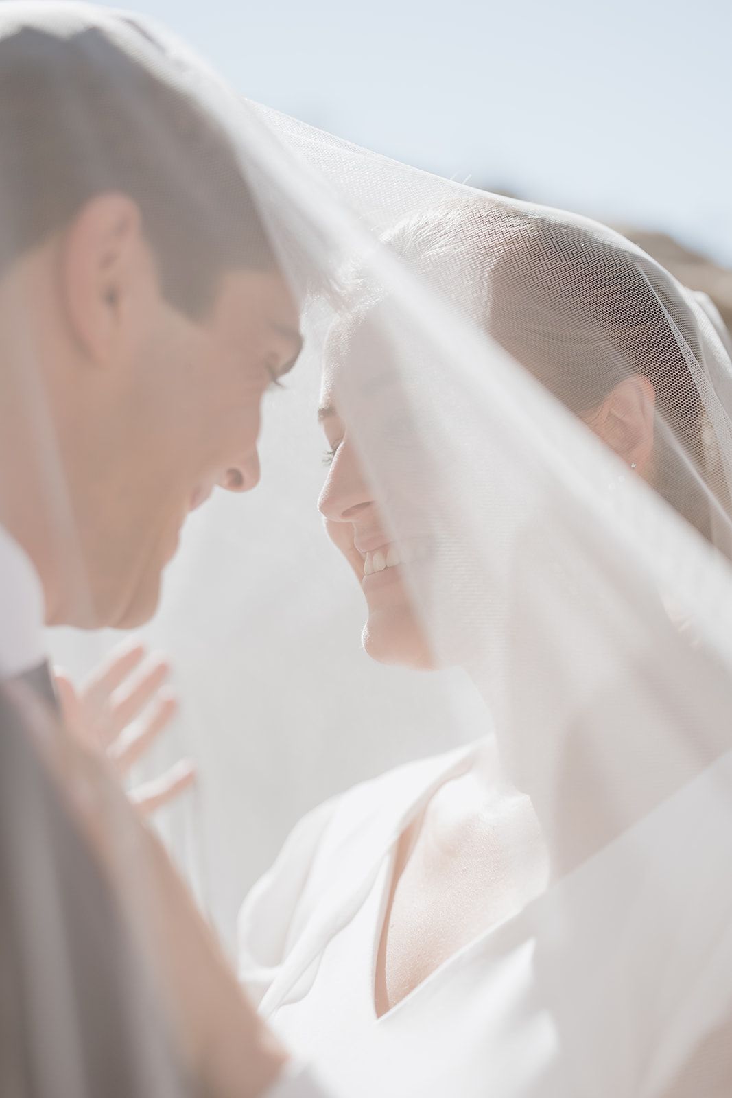Dreamy sun-lit portrait of couple sharing intimate moment beneath sheer wedding veil.