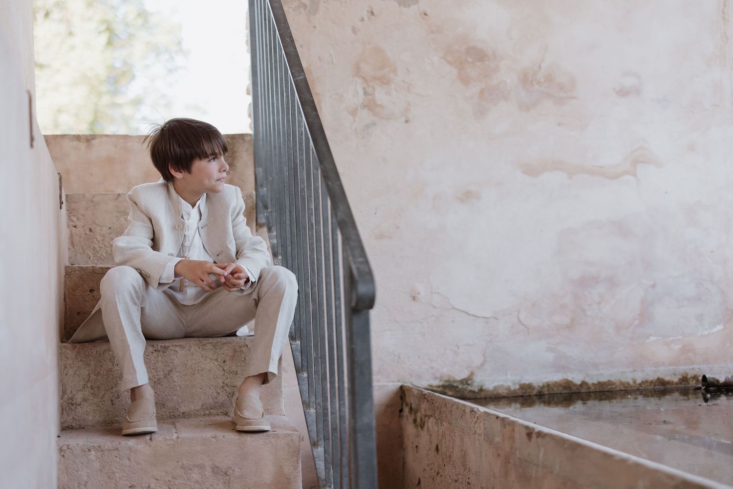 A person in a white suit sits thoughtfully on concrete stairs against a textured wall.