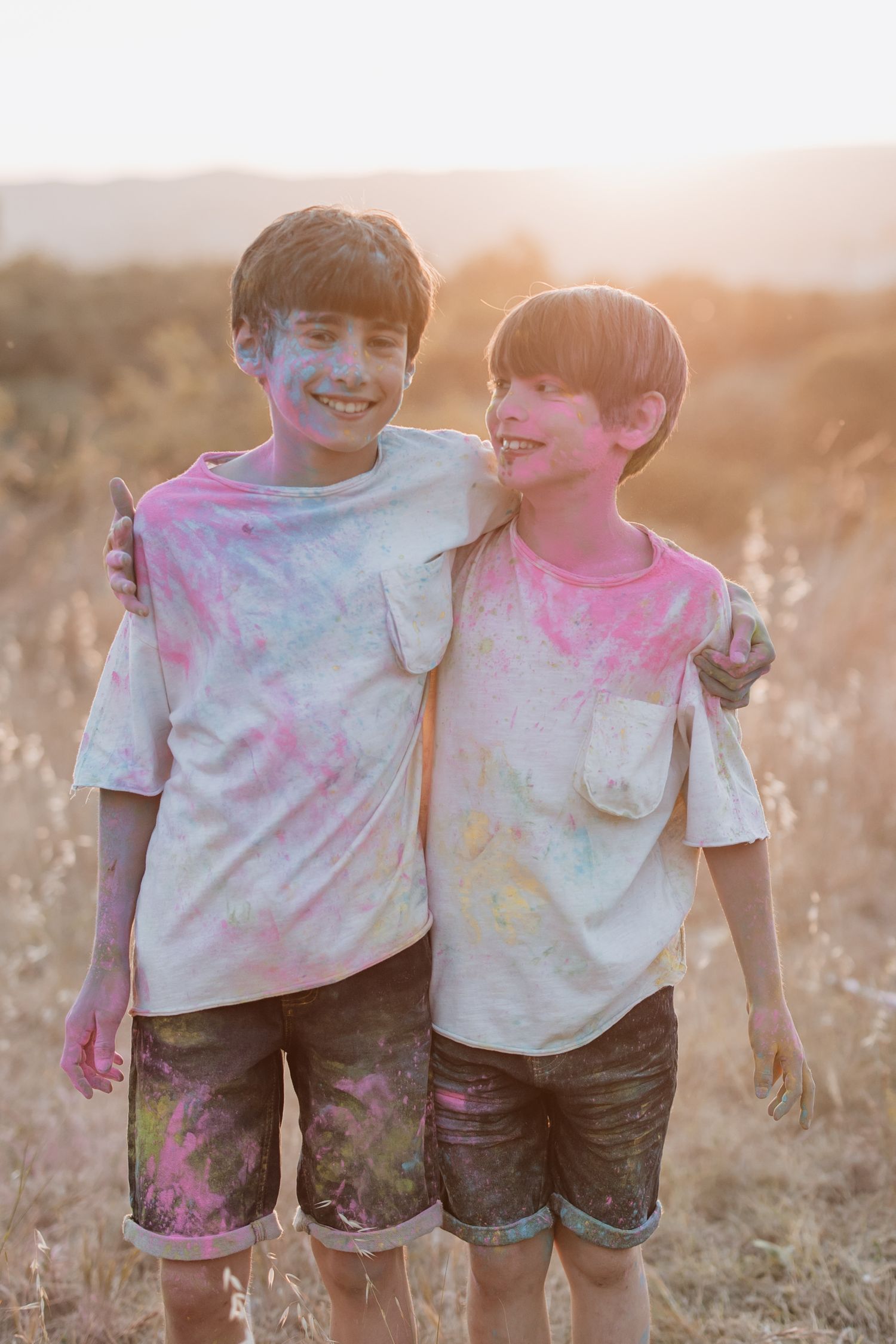 Two figures covered in colorful powder stand together smiling in a sunlit field at golden hour.