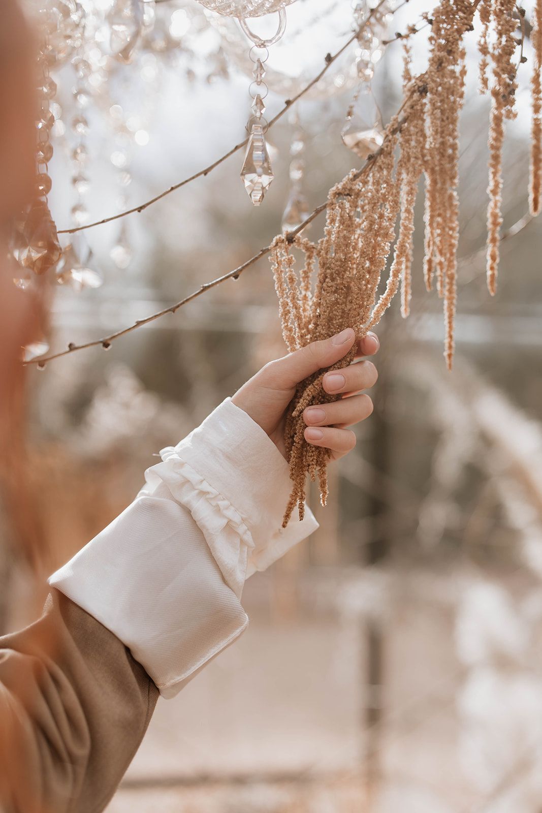 A hand in a white sleeve touches delicate crystal decor hanging from branches in warm lighting.