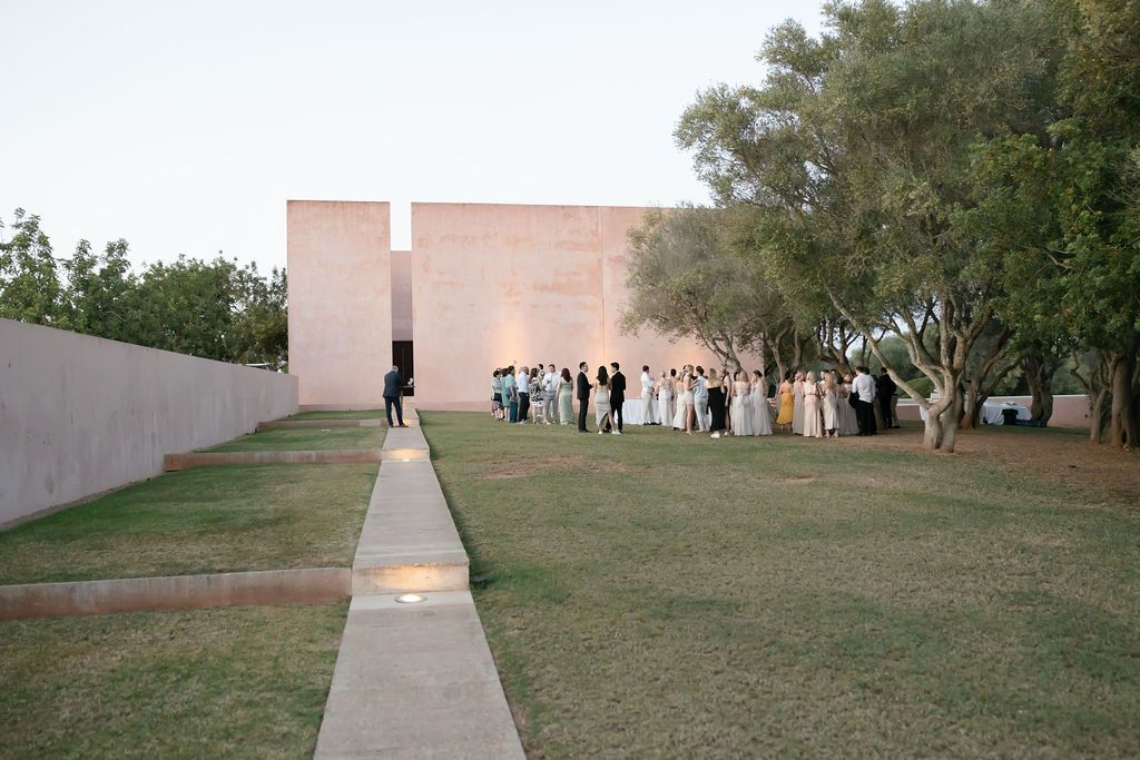 A group of people gathered outside a pink modern building with a concrete walkway and trees.