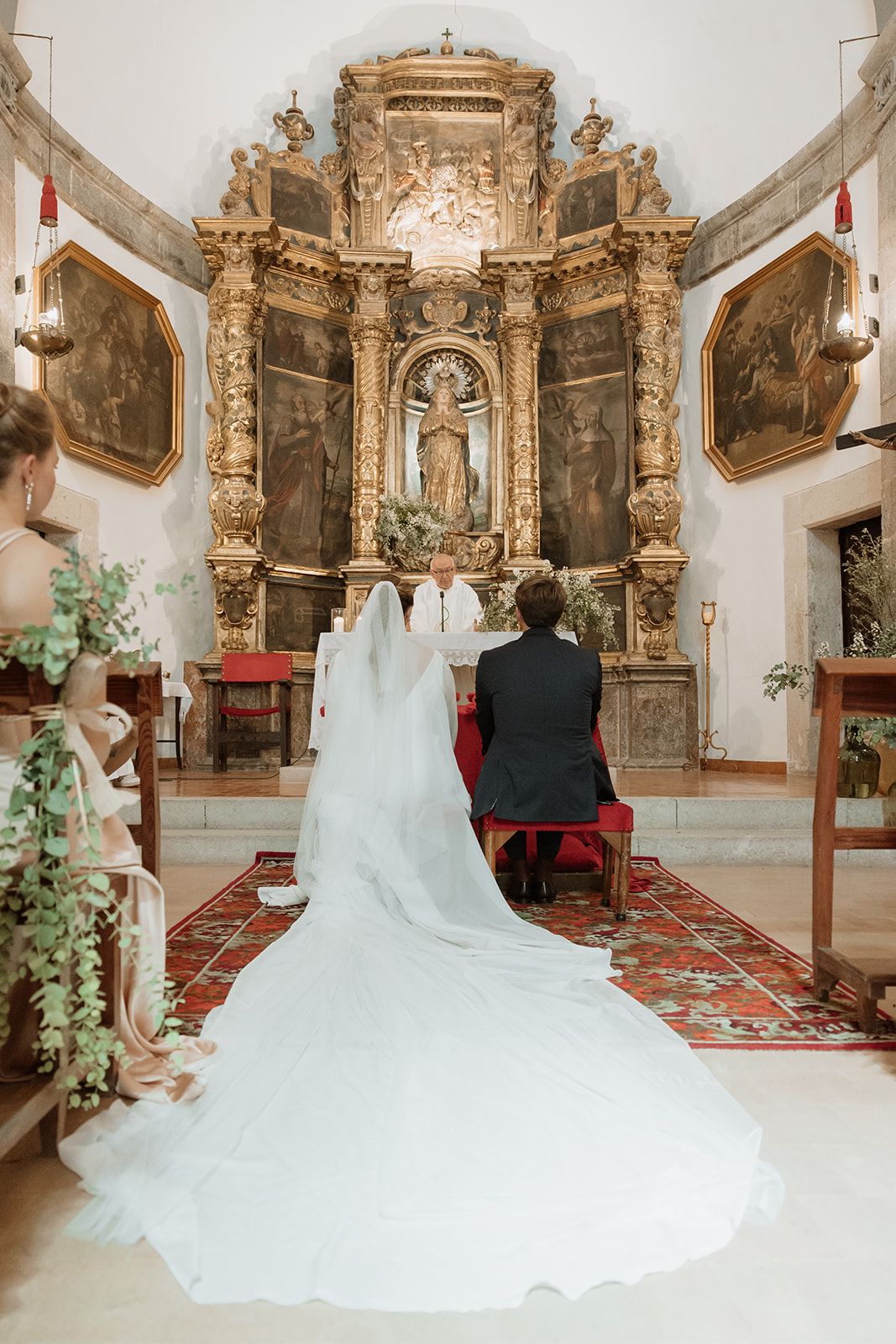 Long flowing wedding dress train extends behind couple kneeling at ornate church altar.