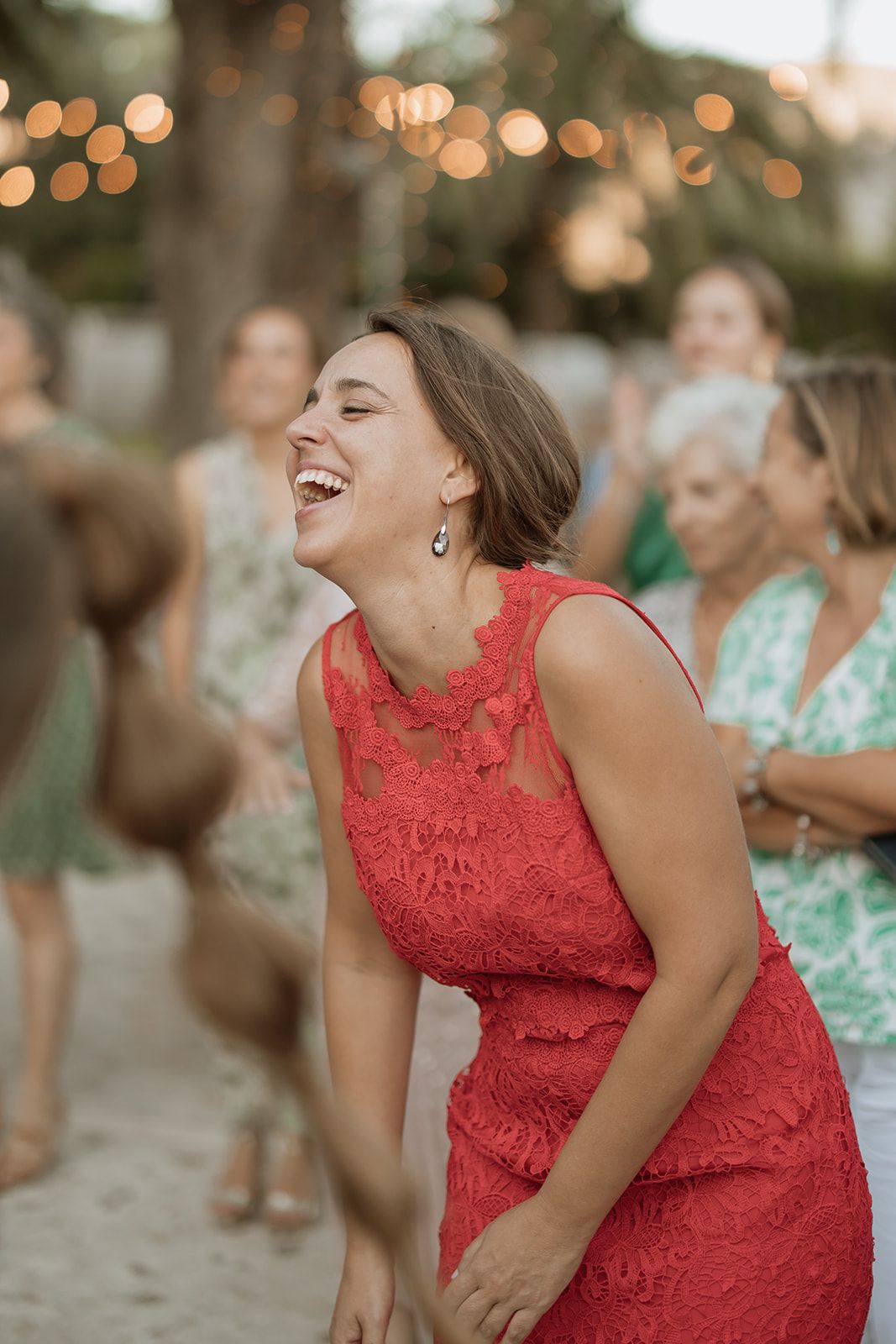 A wedding guest in a coral lace dress laughs heartily at an outdoor reception with string lights.