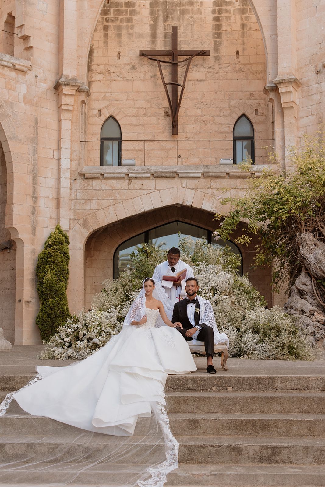 Newlyweds seated on stone steps surrounded by white floral arrangements in historic church courtyard.