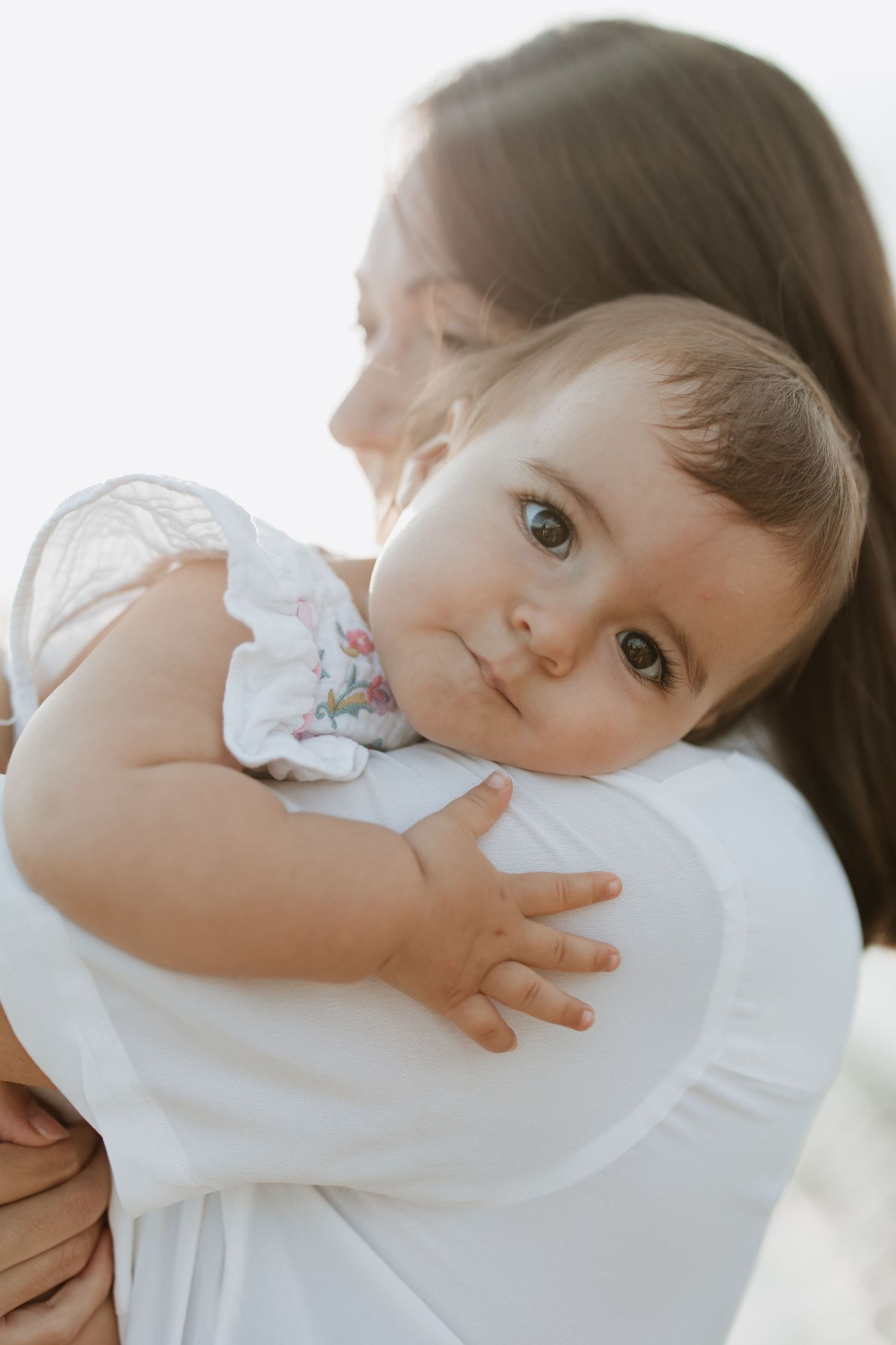 Close-up portrait of a baby in white clothing being held against a white shirt in natural light.