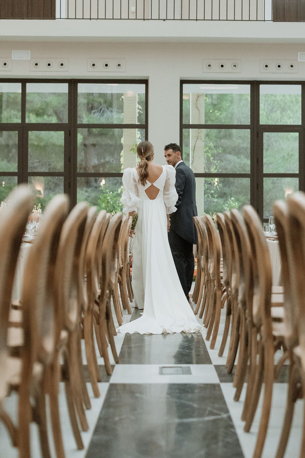 View down aisle of empty chairs towards couple standing in front of tall windows.