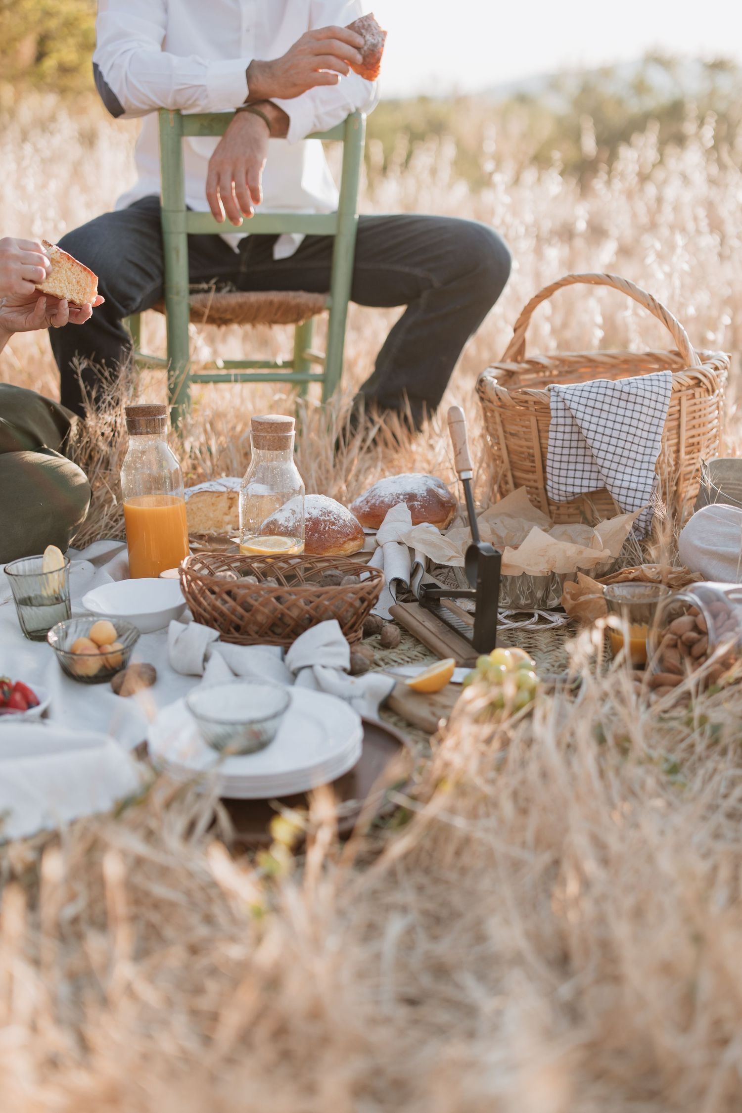 A rustic picnic setup in a wheat field with wicker baskets, fresh bread, and drinks laid out on a blanket at sunset.