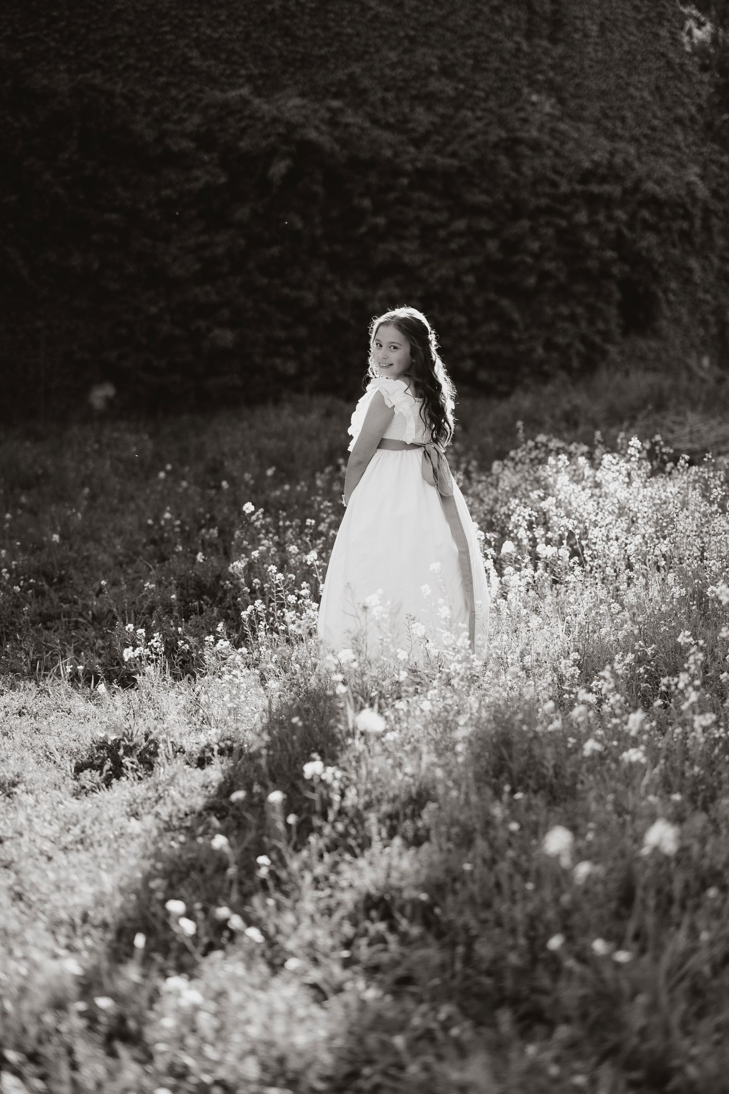 Black and white photo of a girl in a white dress standing in a wildflower meadow, glancing back over her shoulder.