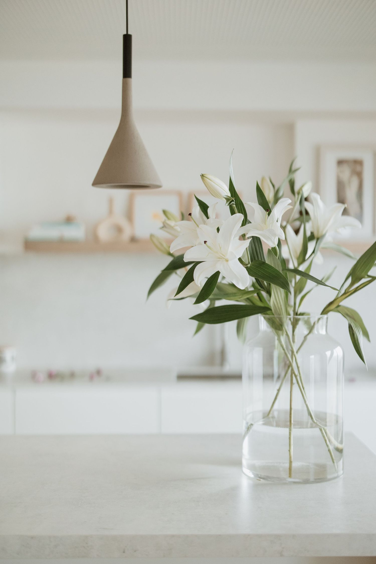 White lilies in a clear vase under pendant lighting on a bright countertop.