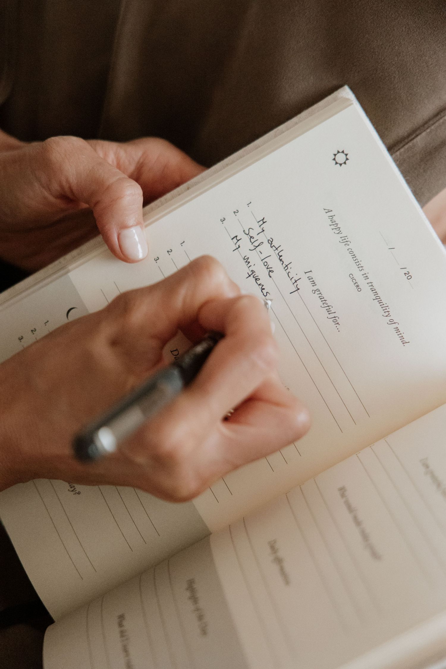 Hands holding pen and writing in an open journal against brown fabric background.