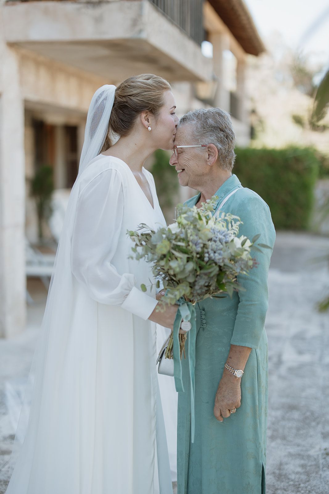 Tender moment between bride in white dress and woman in turquoise dress holding eucalyptus bouquet.